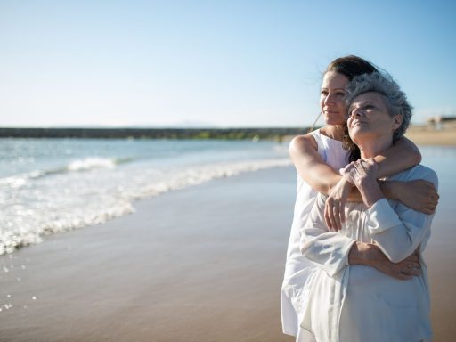 An elderly mother and adult daughter share a warm embrace on a sunny beach in Portugal.
