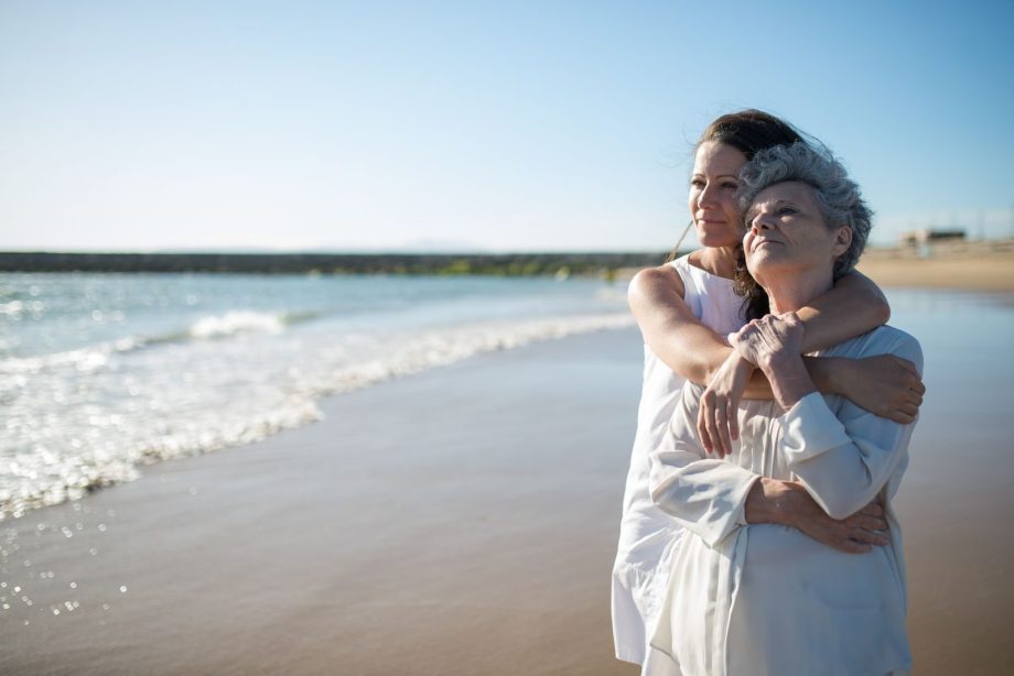 An elderly mother and adult daughter share a warm embrace on a sunny beach in Portugal.